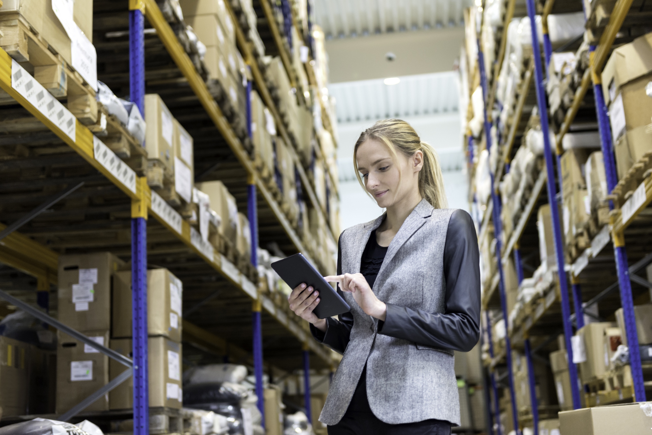 A woman in business attire using a tablet to check inventory in a warehouse aisle, surrounded by tall shelves filled with boxes.
