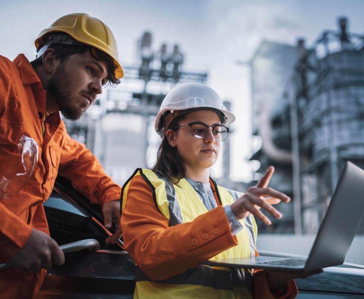 Two workers in reflective vests consult a laptop at an oil refinery, discussing operational data.