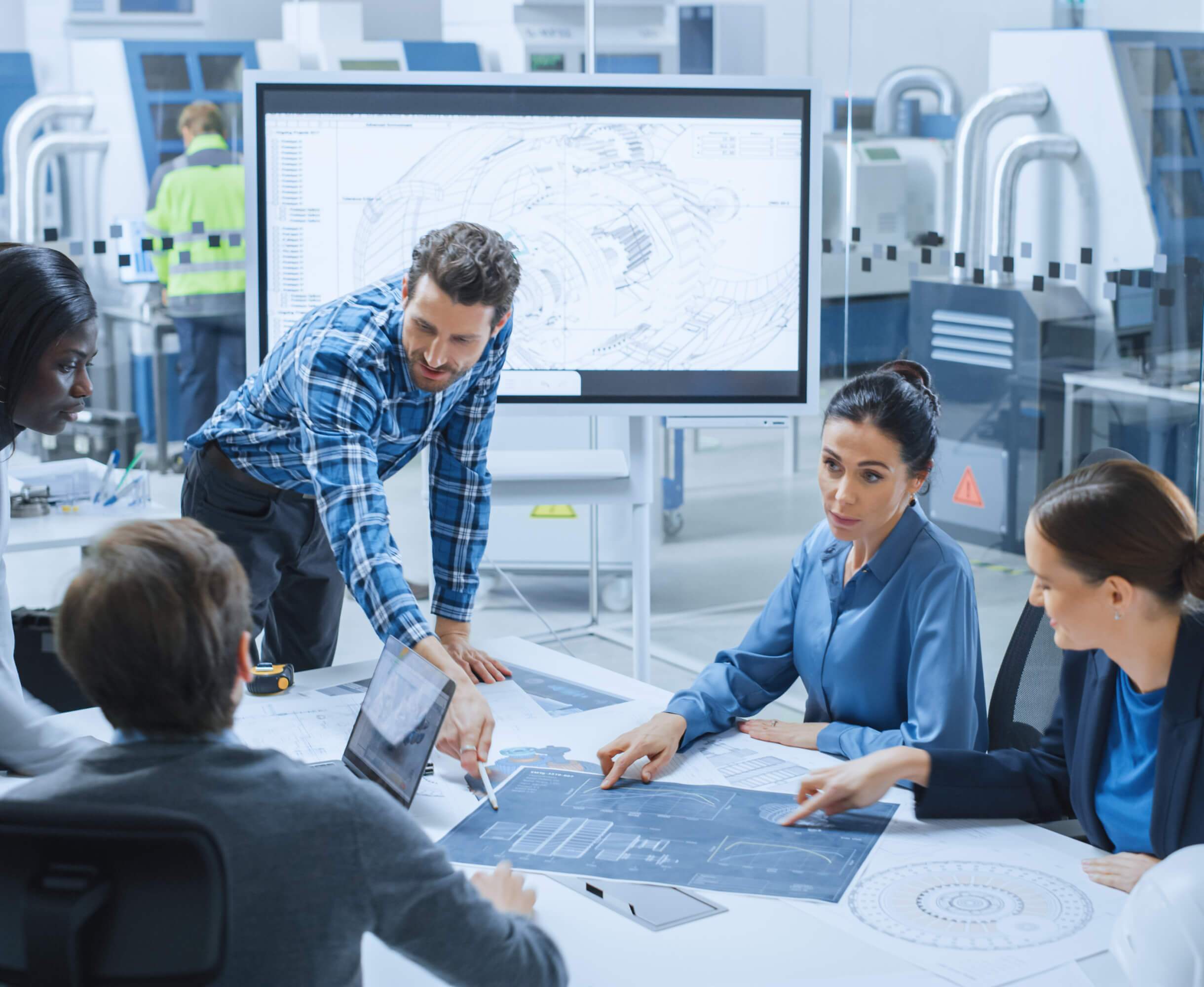 A diverse group of engineers, both male and female, discussing over blueprints in a high-tech manufacturing facility with large digital displays.