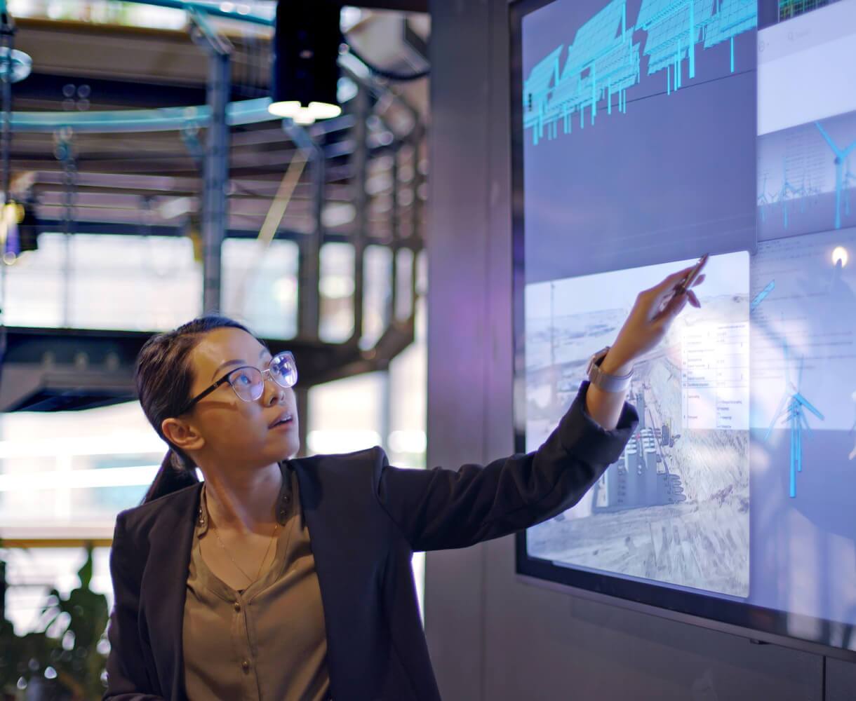 A woman in business attire points to data on a digital display in an industrial setting.