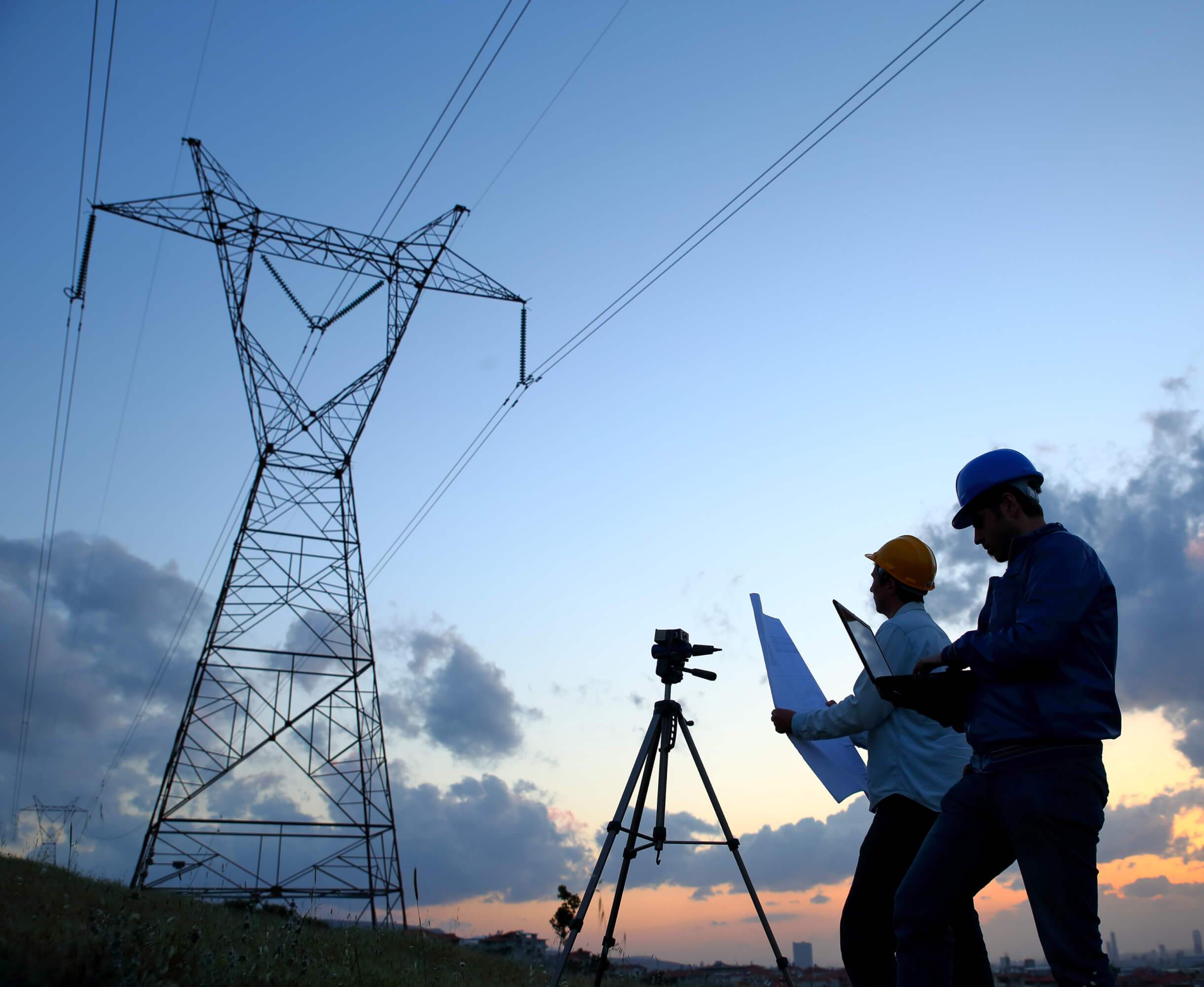 Two engineers reviewing energy infrastructure plans at dusk with a large electricity pylon in the background.