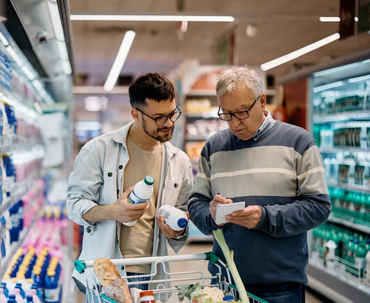Un homme âgé et un jeune homme comparent des produits dans un rayon de supermarché, le jeune homme tenant une bouteille et le plus âgé consultant une liste.