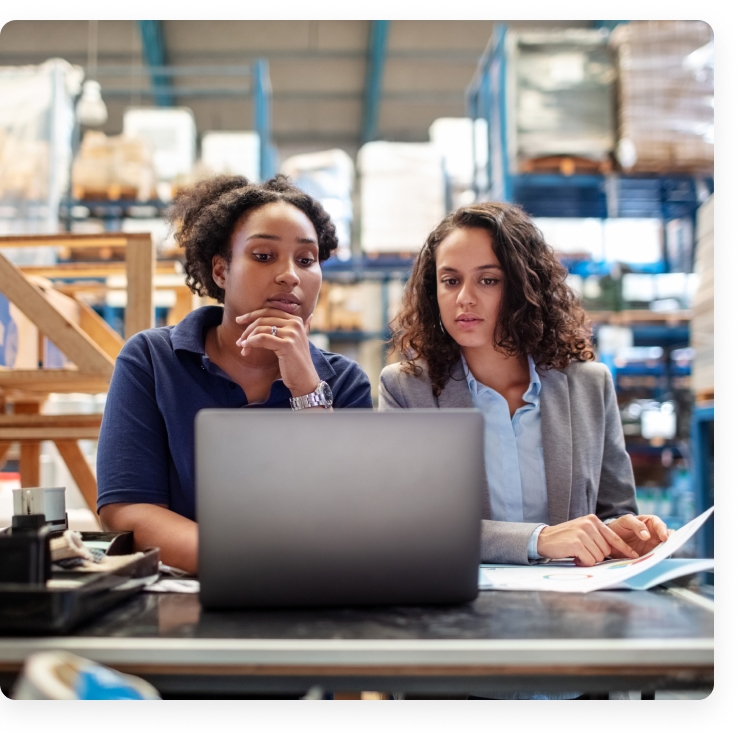 Two female professionals analyzing data on a laptop in a warehouse office setting, focused on the screen.