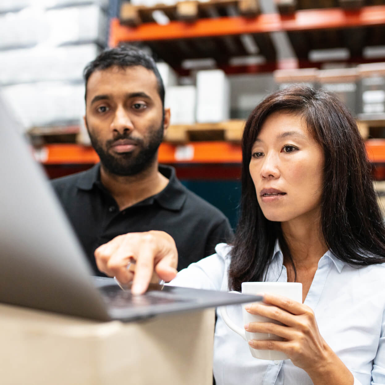 A man and woman, focused and collaborating, view logistics data on a laptop in a warehouse setting.