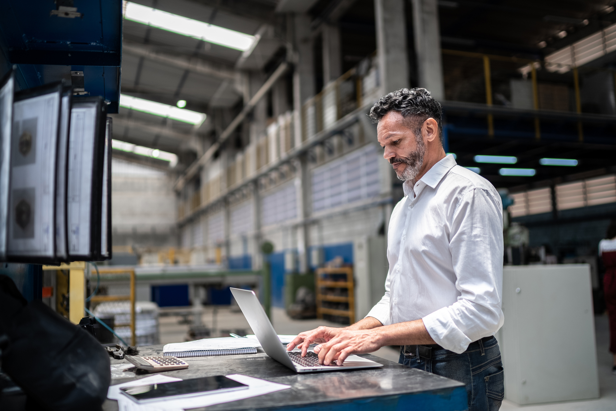 A middle-aged man smiles while using a laptop in a spacious industrial manufacturing area.