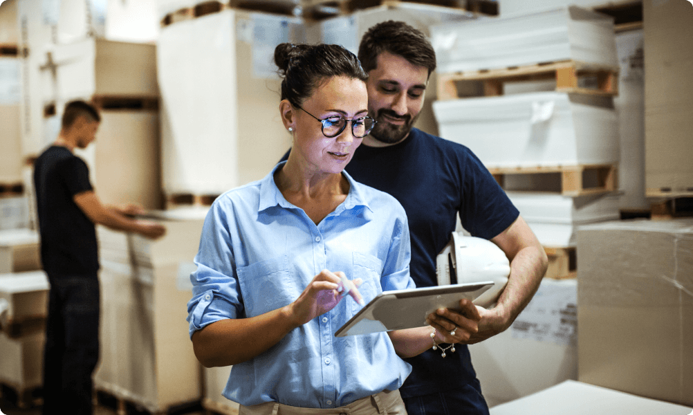 A female manager and a male manager reviewing inventory data on a tablet in a busy warehouse, with another worker in the background.