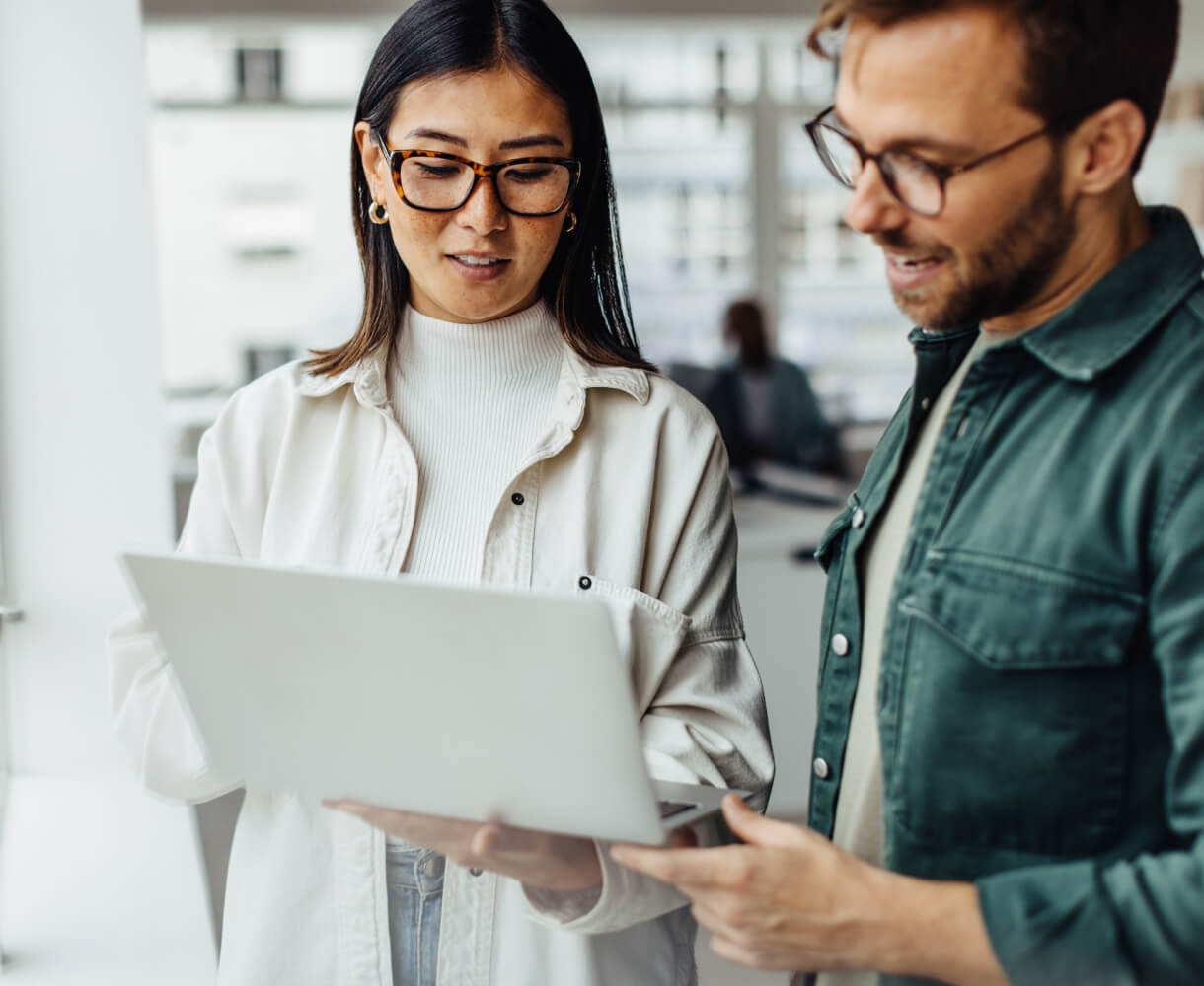 A man and a woman in an office space discuss work-related topics over a laptop.