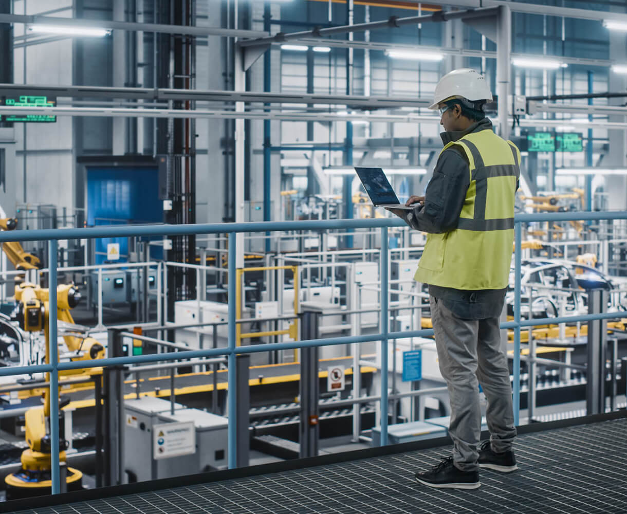 A worker in a hard hat and safety vest uses a laptop on a walkway overlooking a large industrial plant.