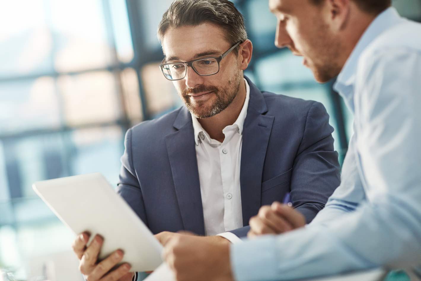 Two businessmen discuss over a tablet in a contemporary office setting.