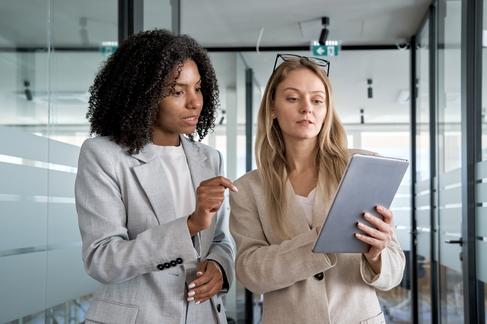 Two professional women, one Black and one Caucasian, intently reviewing data on a tablet in a modern office.