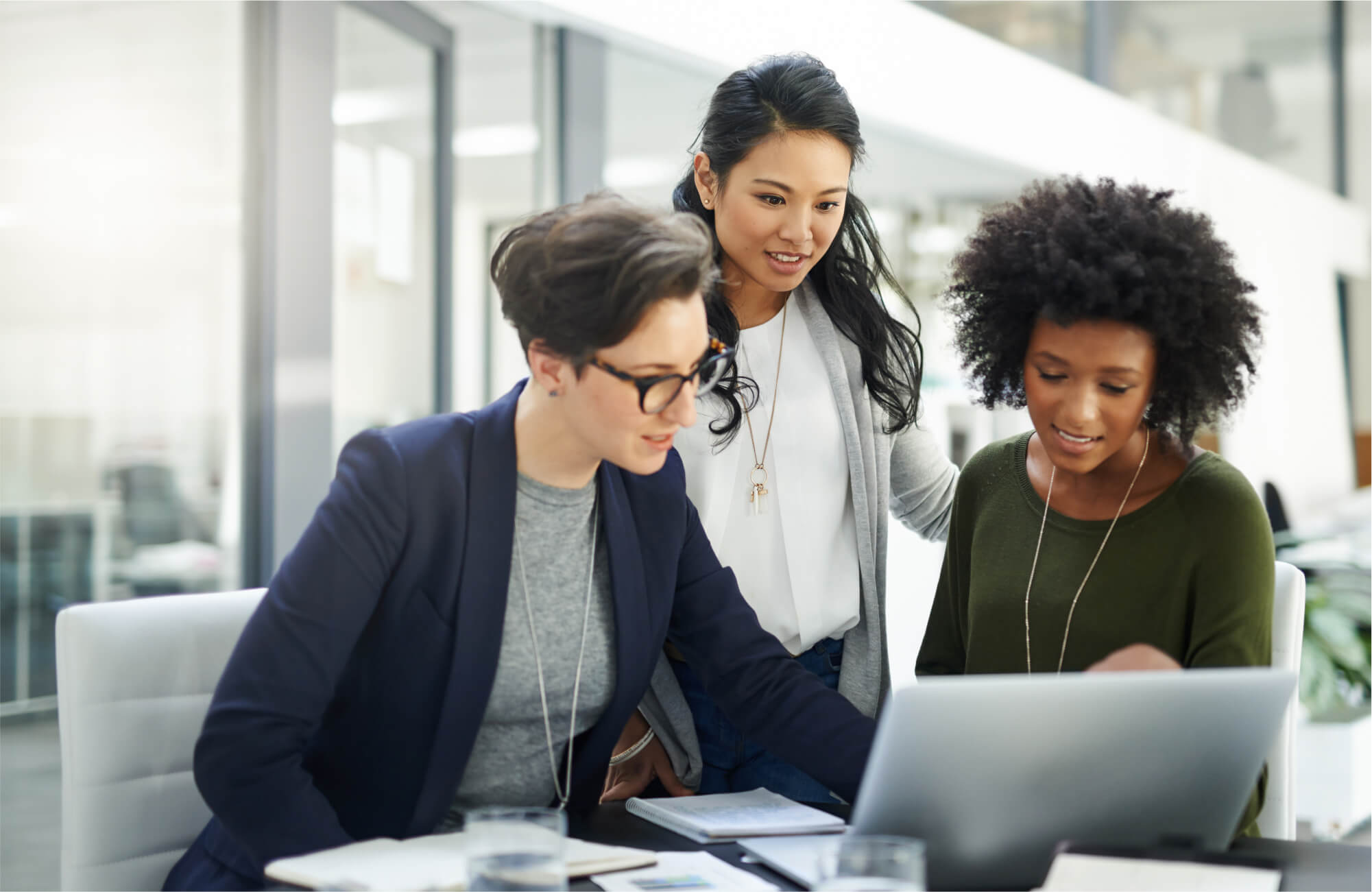 A Caucasian woman, an Asian woman, and an African American woman, working together on a laptop in a modern office setting.